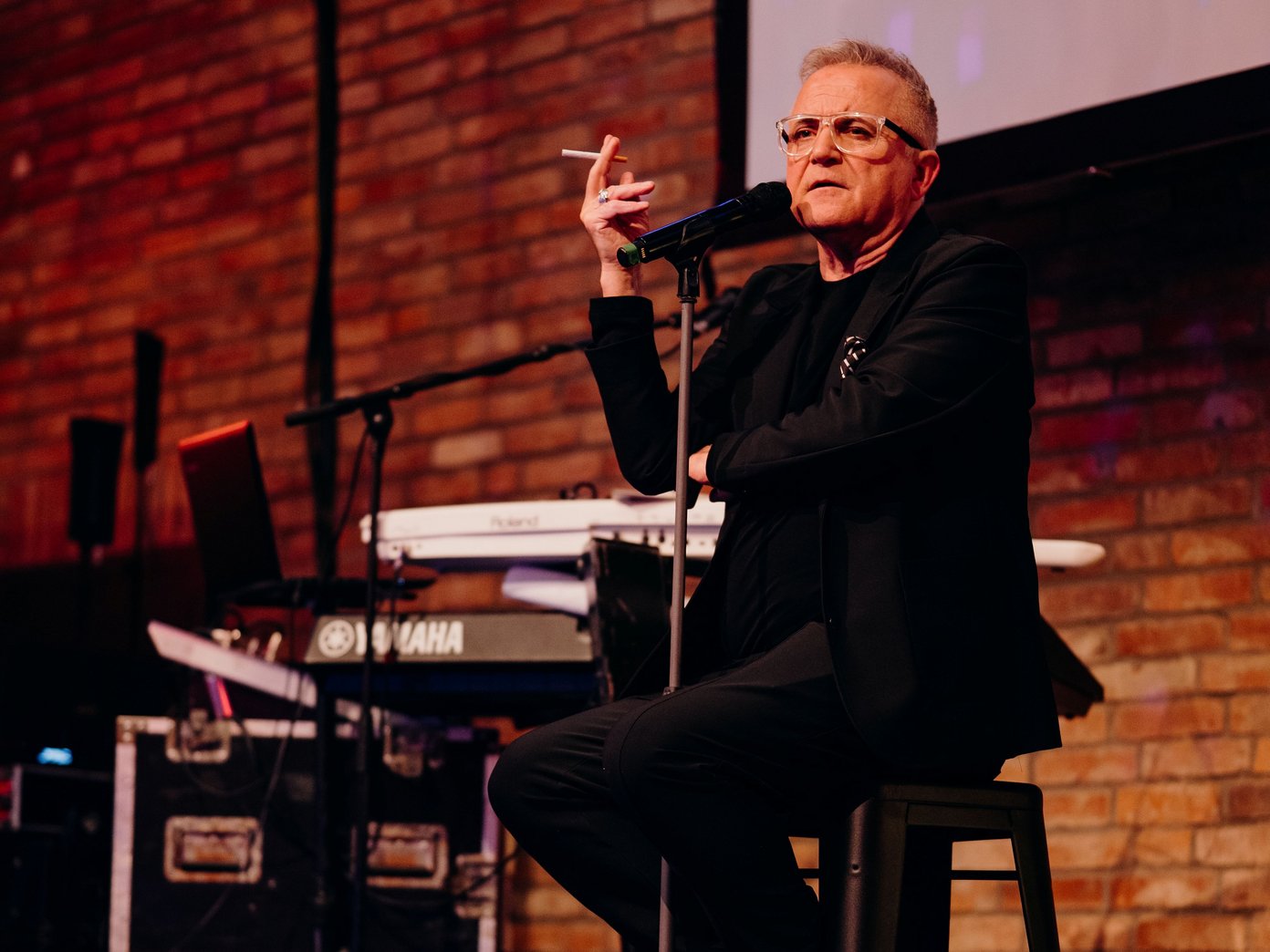 eine männlich aussehende Person mit Zigarrette in der Hand auf einem Hocker am Mikrofon