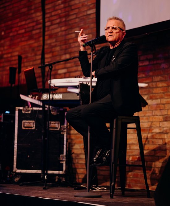 eine männlich aussehende Person mit Zigarrette in der Hand auf einem Hocker am Mikrofon