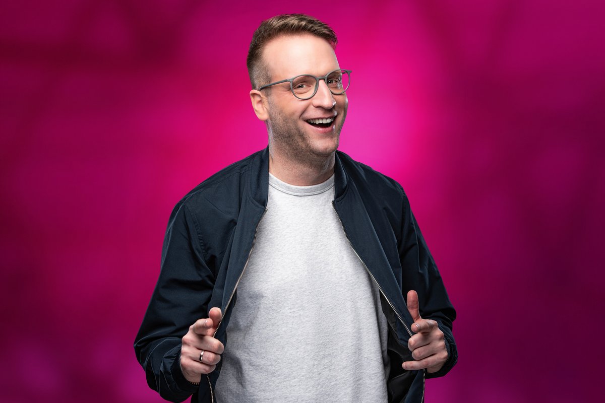 Benny Stark Profilbild Benny Stark zeigt mit zwei Fingern auf das Publikum vor rosanem Hintergrund