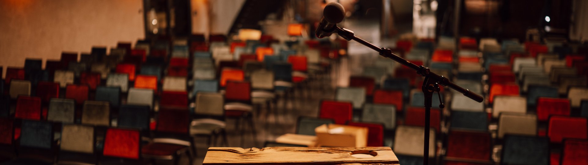 Vordergrund ein Holzpult vor einer Reihenbestuhlung mit bunten Stühlen