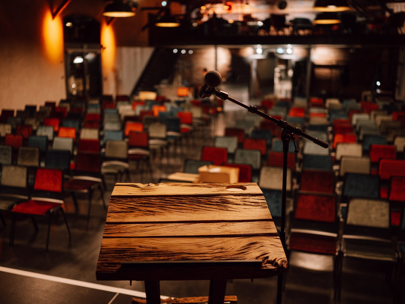Vordergrund ein Holzpult vor einer Reihenbestuhlung mit bunten Stühlen