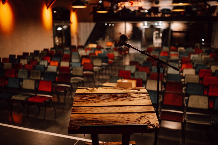 Vordergrund ein Holzpult vor einer Reihenbestuhlung mit bunten Stühlen