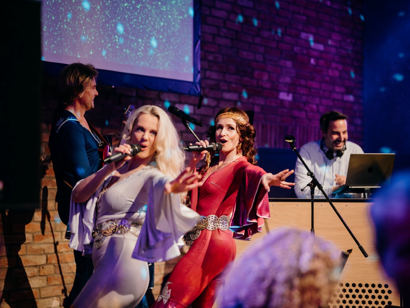 ABBA tanzend zwei weiblich aussehende Personen auf einer Bühe am singen, im Hintergrund ein DJ am DJ- Pult